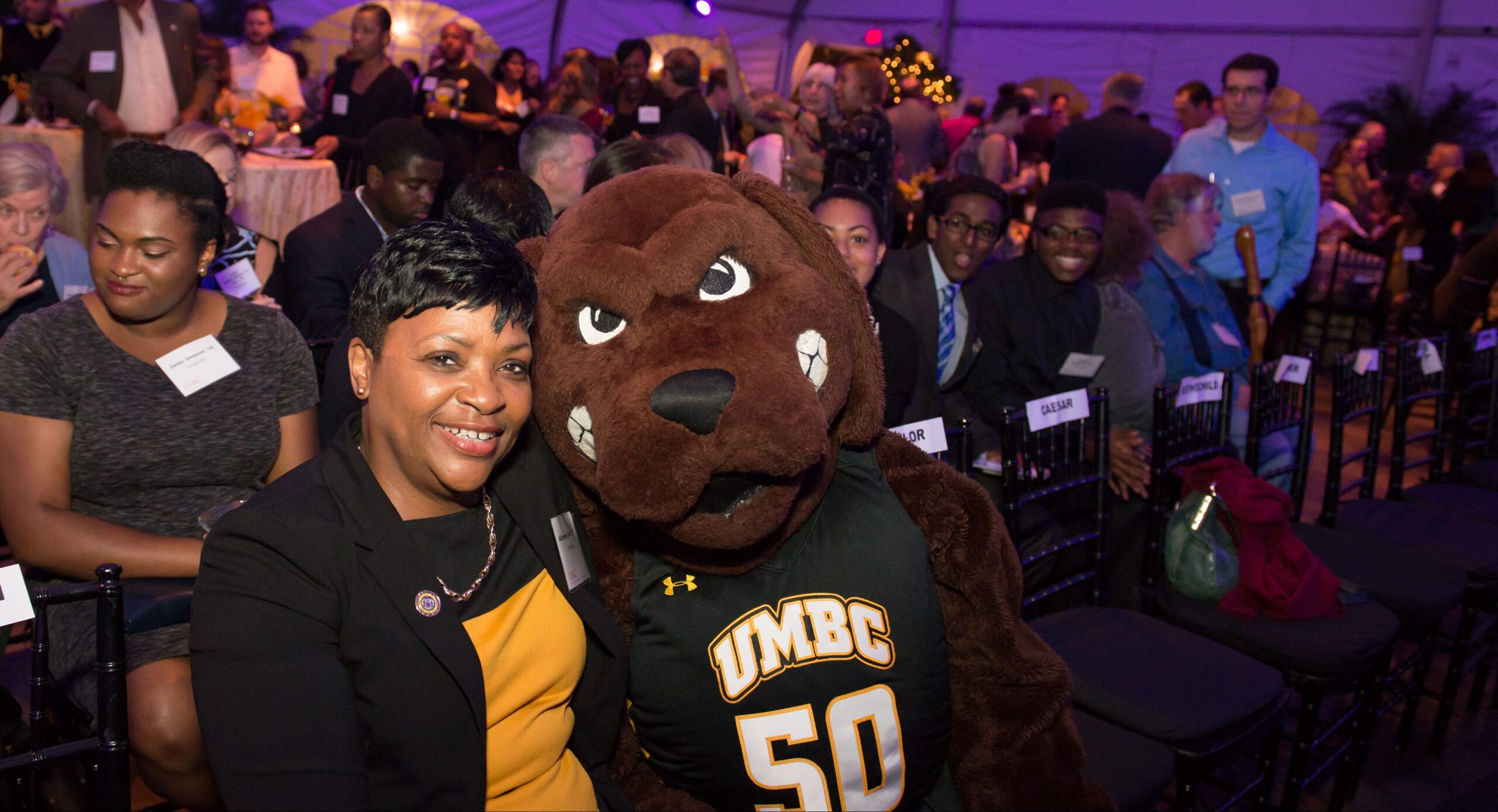 Maryland House Speaker Adrienne Jones, in gold and black, poses with UMBC Retriever mascot, in front of a crowd of people, during an evening celebration.
