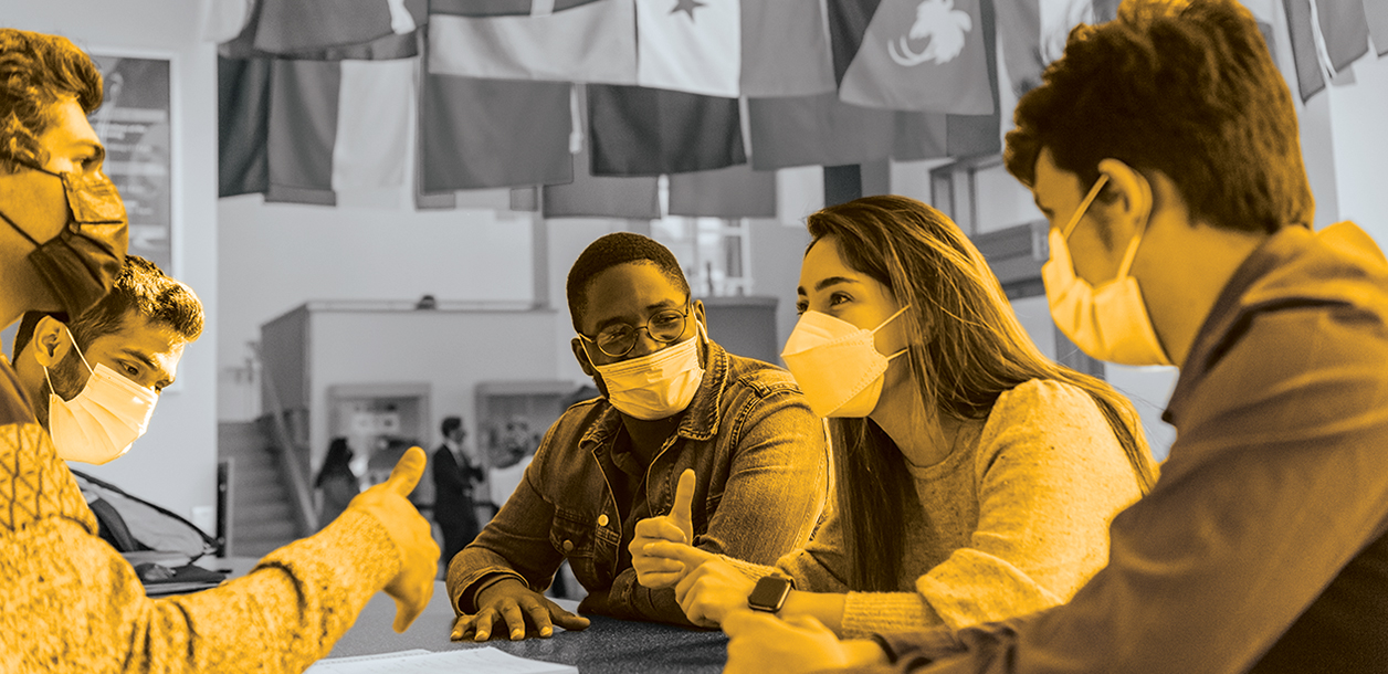 In gold overlay, students sit around a table in the commons talking.