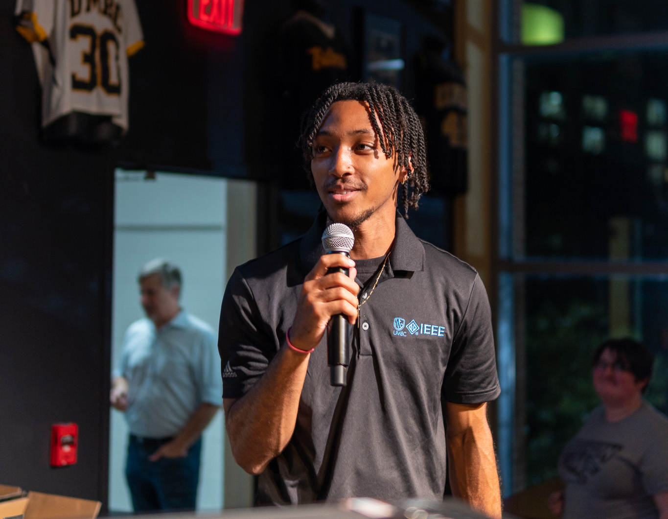A young man stands in a black polo shirt talking into a microphone