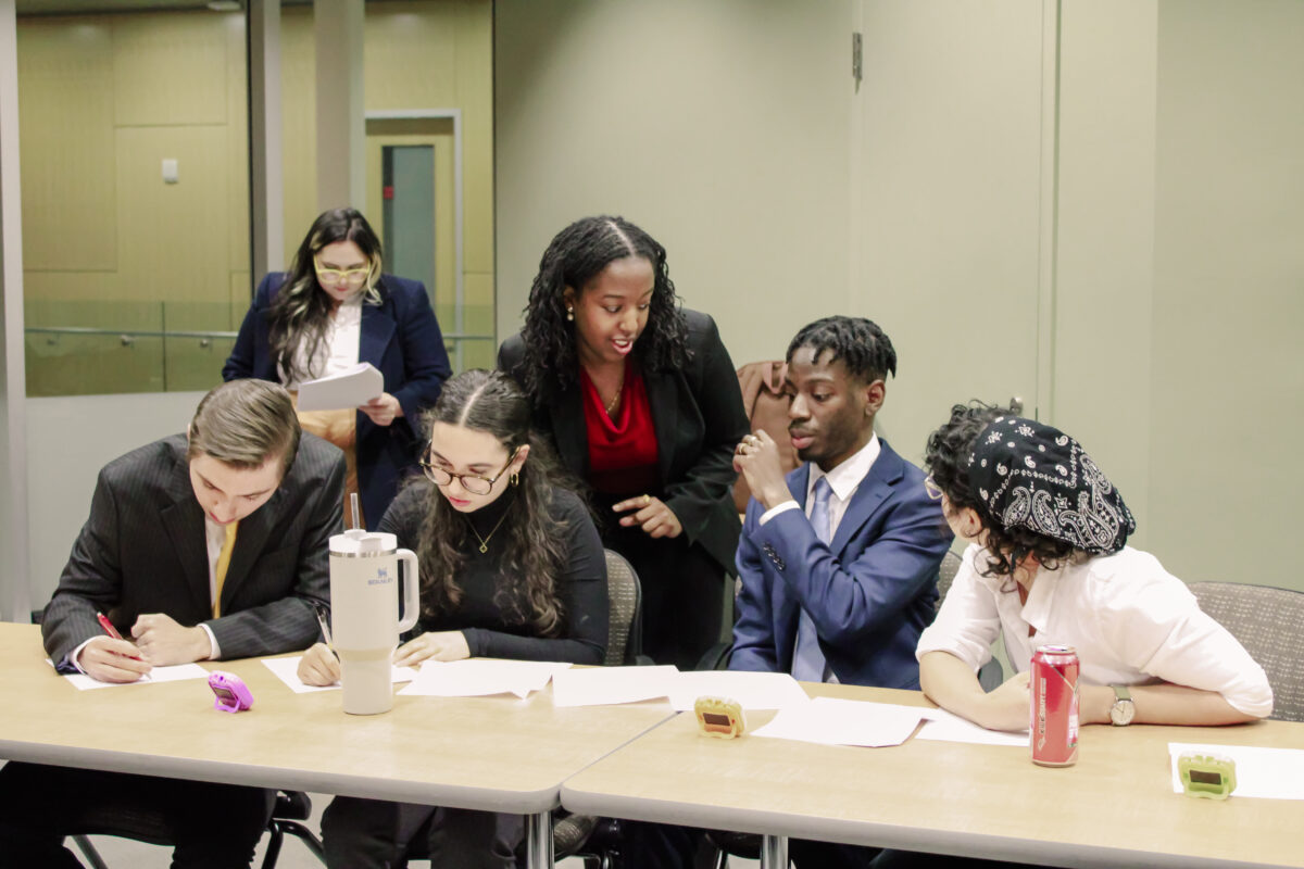 Five college students dressed in formal wear sit at a table discussing and taking notes