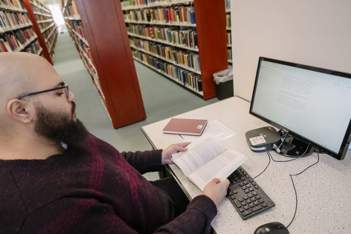 A humanities college student sits at a desk looking through a book in front of a computer screen open to a text document