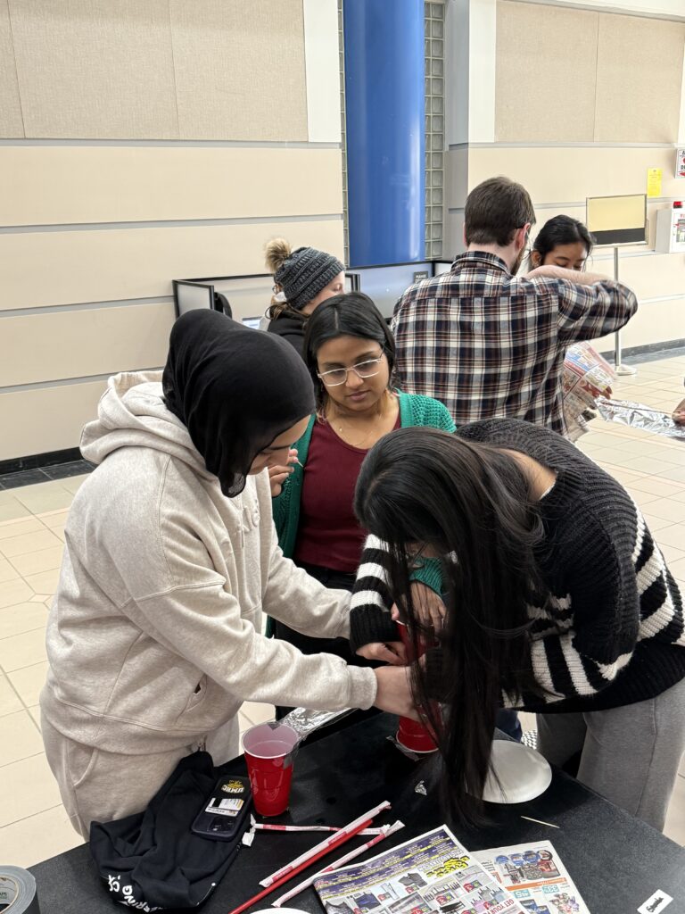 Students work together around a table, building something with plastic cups, tin foil, straws.